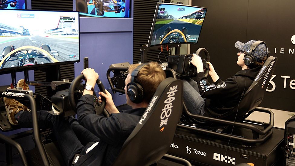 Two players race on an F1 game. They're sitting in gaming chairs with steering wheels and pedals designed to mimic the cockpit layout of a Formula One car. They're concentrating intensely on two large screens attached to the front of the gaming rigs.