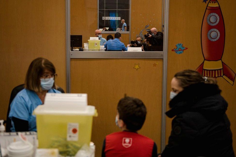 A child sits with his mother as he waits for his dose of the Pfizer-BioNTech vaccine for children as photographers photograph another child receiving the vaccine in November 2021 in Montreal, Quebec