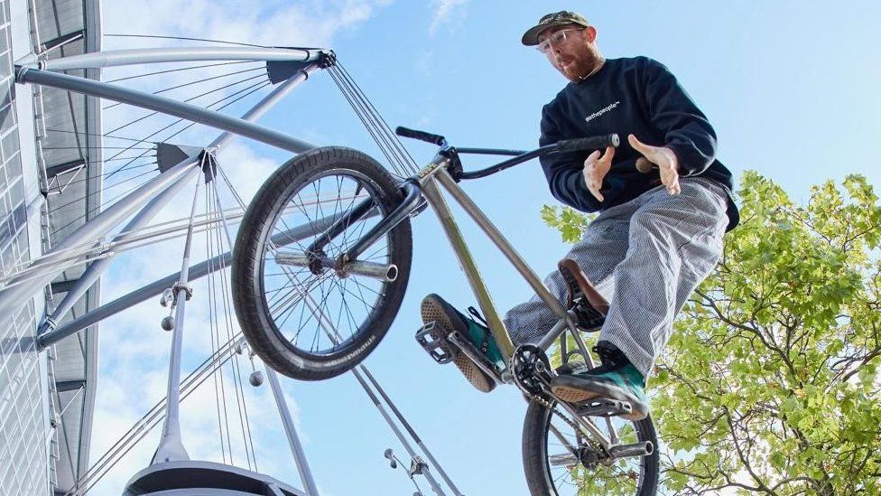 Dan Banks, a white man in his late 20s, pictured jumping on his BMX bike. Dan is wearing a green cap, clear square-framed glasses, a dark blue sweatshirt, denim jeans and green trainers. He has a short reddish-brown beard and is looking down at his moss green bike which is in the air above the photographer, his tongue sticking out in concentration. His feet are on the pedals but his hands are not on the handle bars. He's pictured outside on a sunny, blue-sky day with a tree and bridge behind him.
