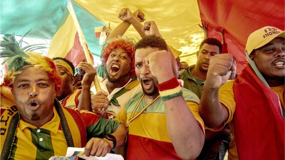 A group of Ethiopian men cheering and shouting. Some of their fists are in the air. They are wearing the colours of the Ethiopian flag.