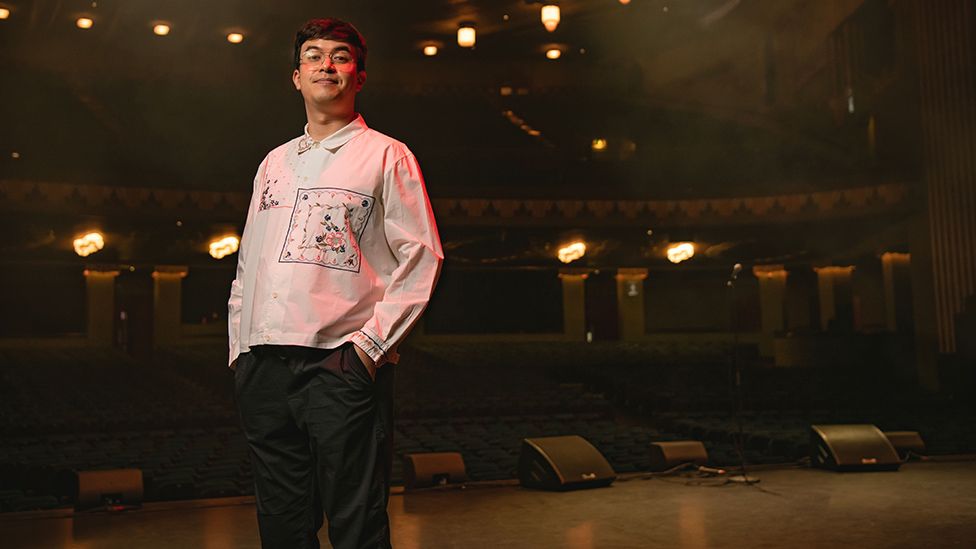 A man with short black hair and glasses stands on a stage as a darkened theatre auditorium stretches out behind him. He's wearing a white shirt with a large, square embroidered floral pattern over his left sternum. He's got his hands in his pockets and looks relaxed.