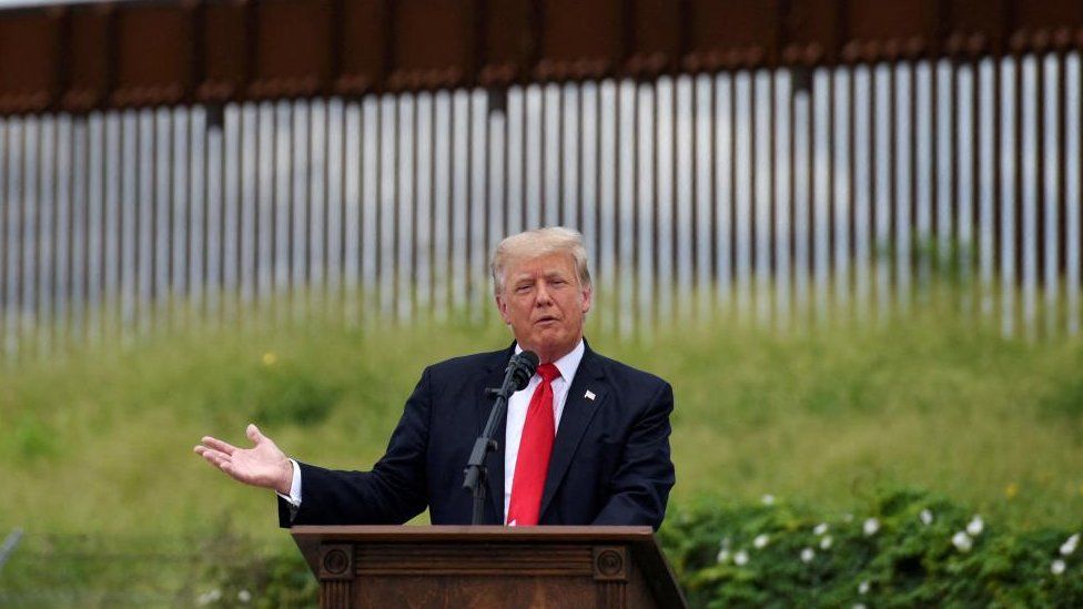 Donald Trump speaks at a lectern, with a section of border wall behind him