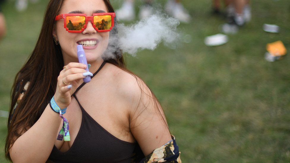 A young woman vapes at Reading festival. She's holding a lilac disposable vape, exhaling vapour. She's wearing a black bikini top and smiling