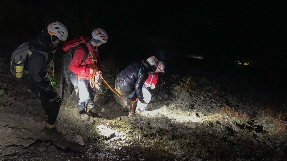 Two women wearing helmets and harnesses are helped down a rocky slope by two mountain rescue teams using ropes in the dark