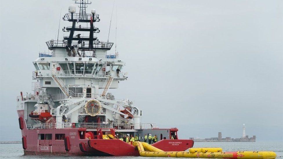 Red and white ship towing a yellow inflatable boom