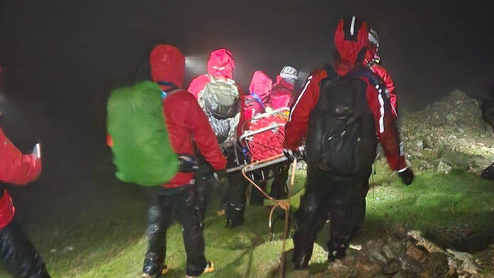 Rescuers in red coats carry a shelter down a steep slope in the dark and rain