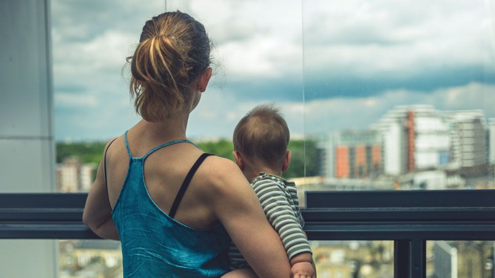 Mother and baby looking out of their apartment at city beyond