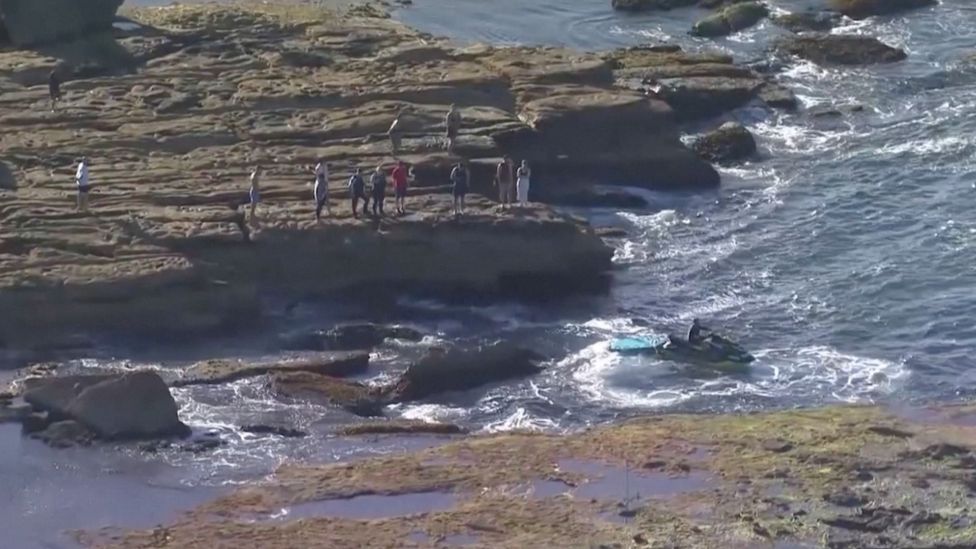 Onlookers and emergency workers stand on the rocks near where the swimmer was found on Wednesday