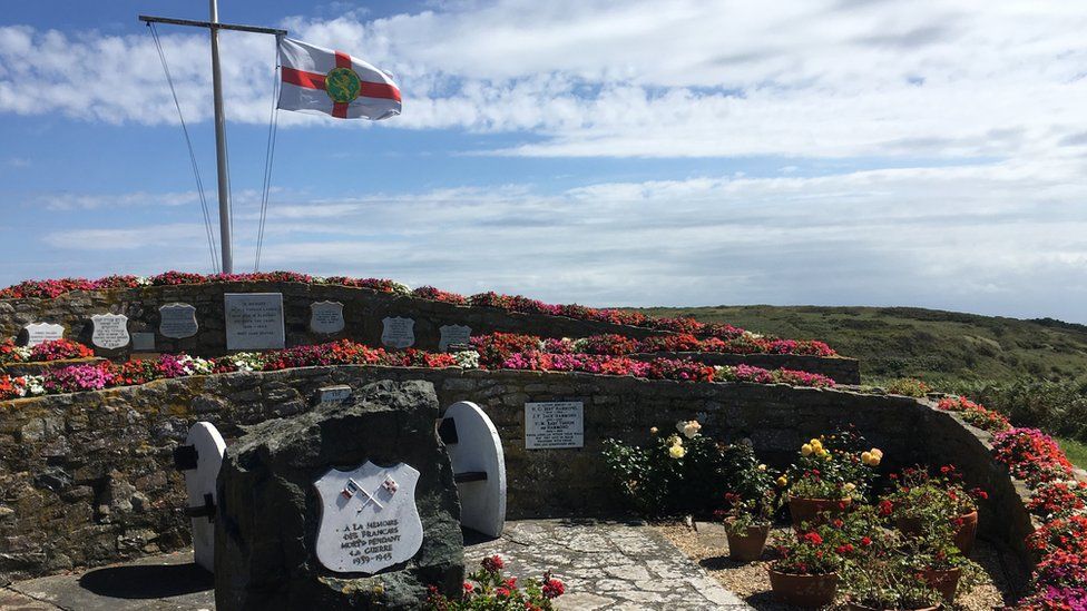 Alderney's Hammond Memorial with flowers in bloom and an Alderney flag fluttering in the wind