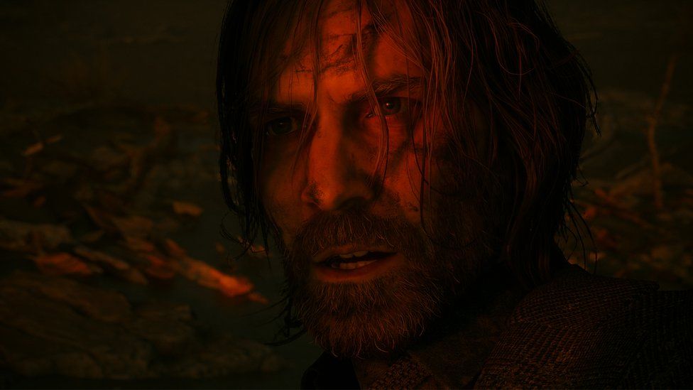 Alan Wake, the title character, seen in footage from the sequel to the first game. He's mostly in darkness, but bathed in a faint, red light. It creates a sinister feeling, and Alan himself looks dishevelled, his long hair covers much of his face and he appears to be covered in mud, blood or a mixture of both.