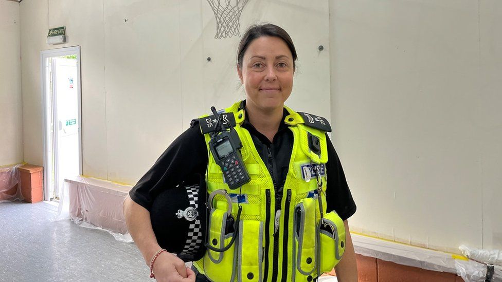Officer Gemma Rutter smiling at the camera. She has dark hair and is wearing a hi-vis police vest with a radio attached to it. She is standing in the redecorated scout hall