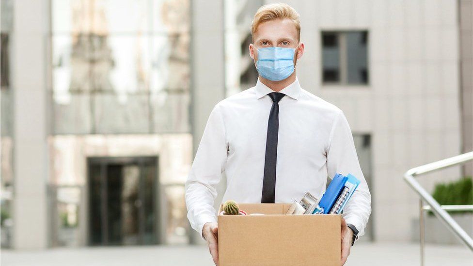 Stock image of a man in facemask carrying a box of his belongings from the office