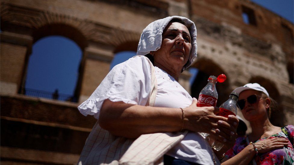 A woman queues to fill bottles with water in Rome as she shelters from the sun with a towel