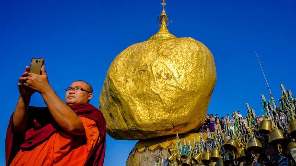 Buddhist monk takes a selfie in front of a gold rock
