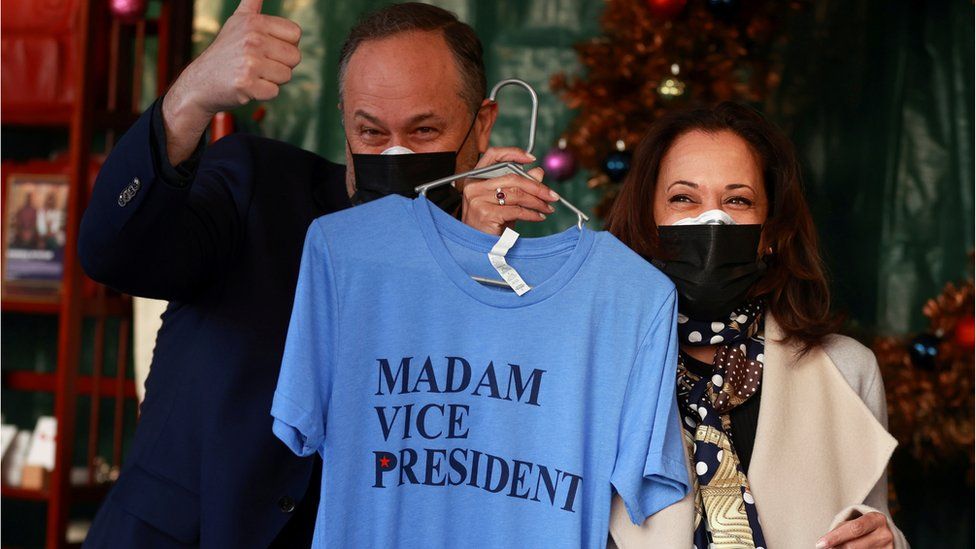 Kamala Harris holds a t-shirt that reads Madam Vice President, while standing next to her husband Doug Emhoff