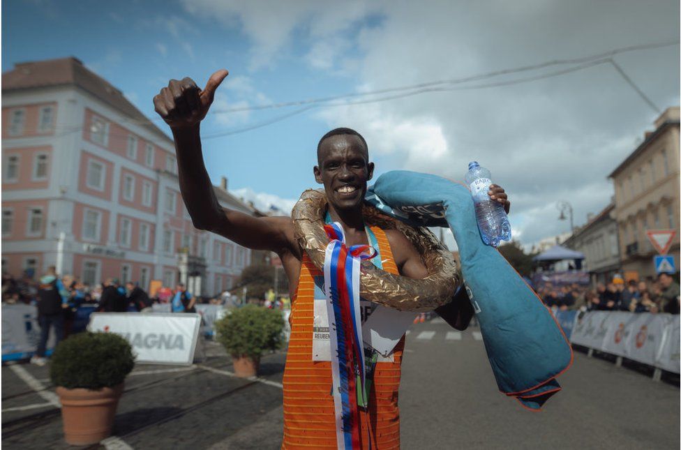 Reuben Kiprop Keiro from Kenya making a thumbs up sign with a bottle of water in hand. He is smiling in an orange singlet against a backdrop of buildings.