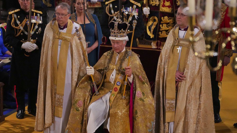 King Charles III is sit at Westminster Abbey wearing a golden cape and St Edward's crown. He is holding sceptres in both hands.