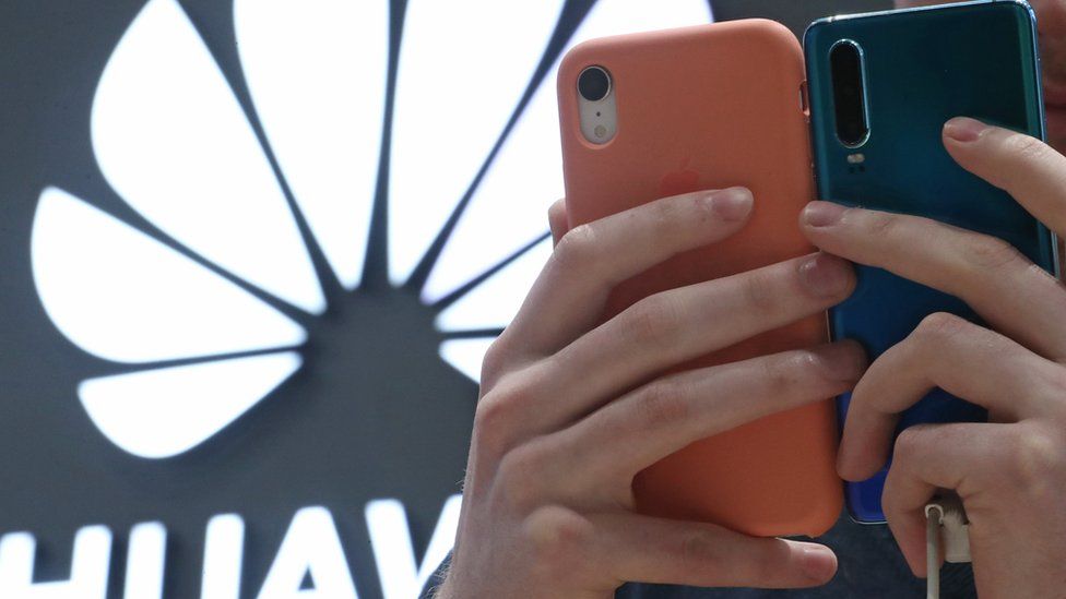 The Huawei logo is seen behind a man's hands, holding two Huawei phones