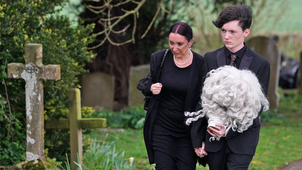 Daughter of Paul O'Grady, Sharyn Mousley, with an unidentified young man carrying holding the wig of Lily Savage arriving for the funeral of Paul O'Grady at St Rumwold's Church in Aldington, Kent.