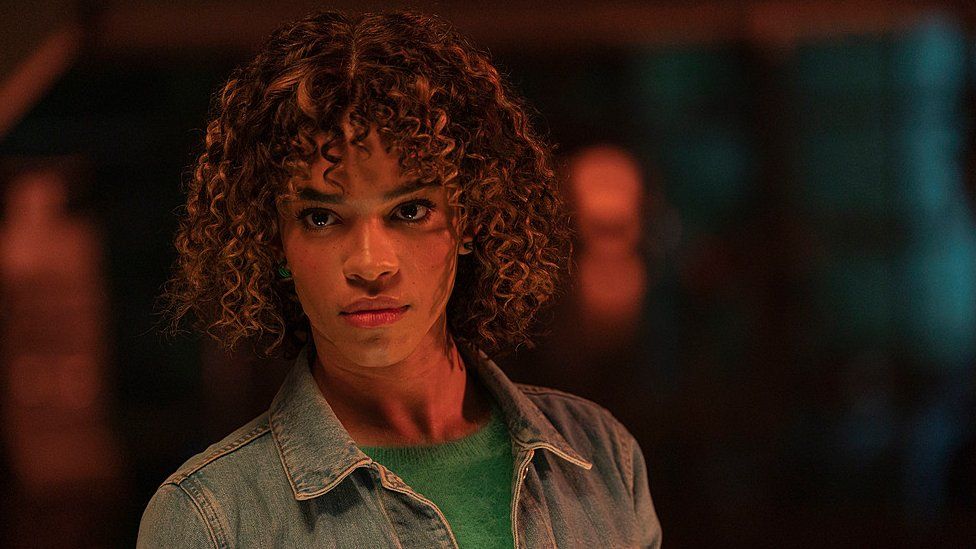 Yasmin Finney, 20, as Rose in Doctor Who. Rose has short curly hair and brown eyes. She wears a denim jacket over a green jumper. She has a serious expression and is pictured inside in dark surroundings.