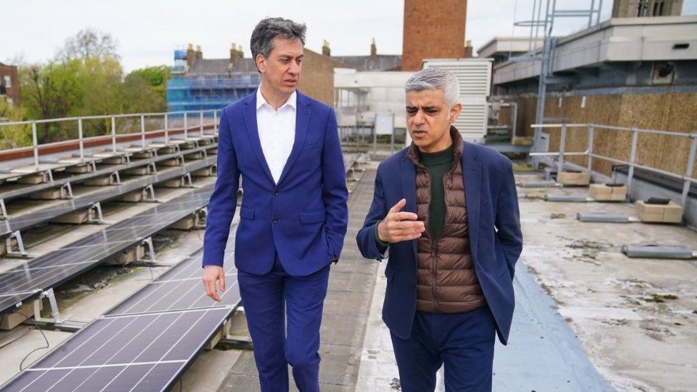 Ed Miliband listening to Sadiq Khan talking as they walk on the roof of a building with solar panels