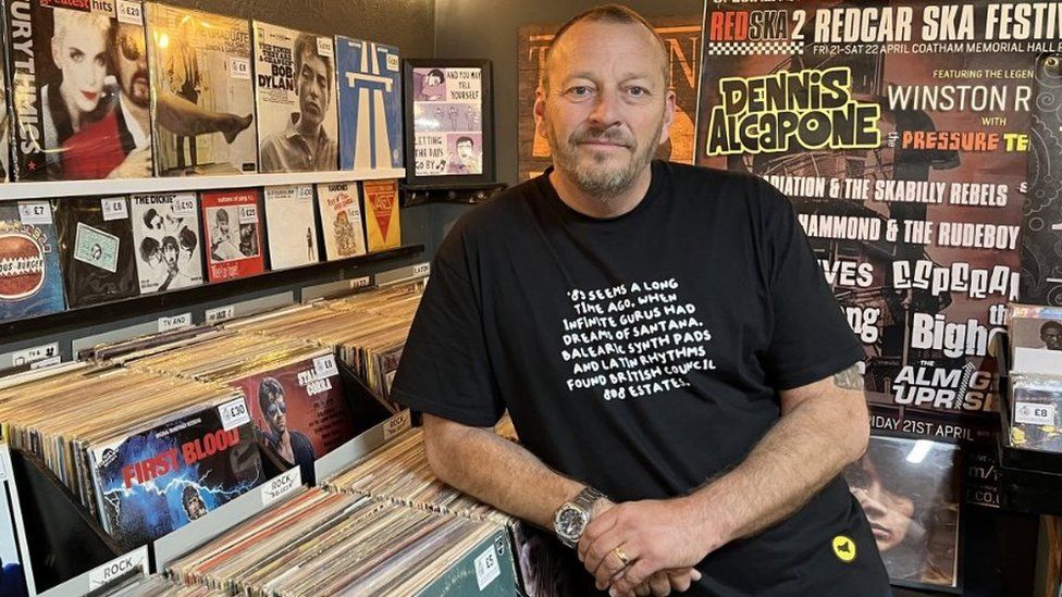 A man in a black T-shirt leans on a rack of records