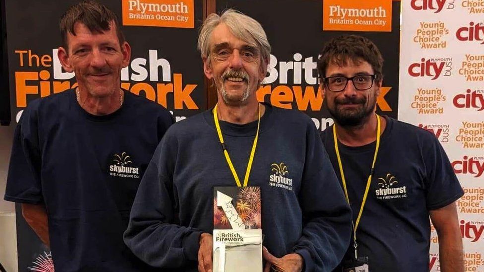 Three male employees from Skyburst. Alan Christie is stood in the middle, holding an award for the British Firework Championships. He has grey hair and facial hair, and is wearing a navy blue sweatshirt with a Skyburst logo on the chest.