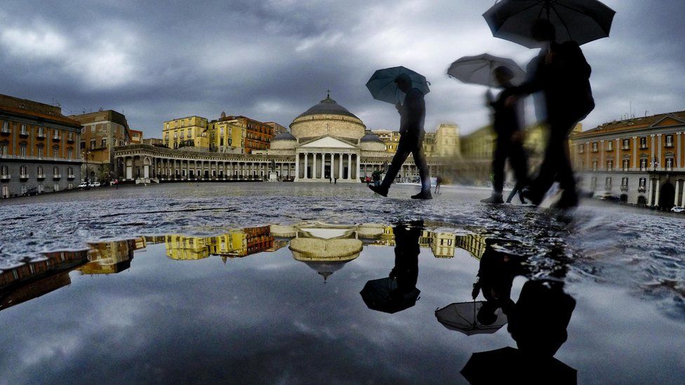 Pedestrians are reflected in a puddle as they protect themselves from the pouring rain with umbrellas while walking in downtown Naples, southern Italy