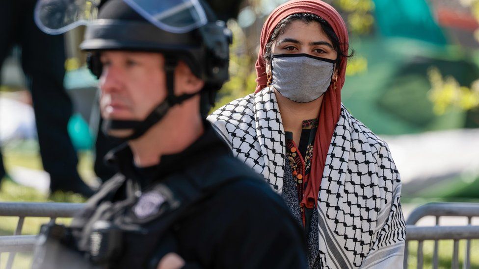 Police watch over protesters at Northeastern University and a woman stands behind the officer