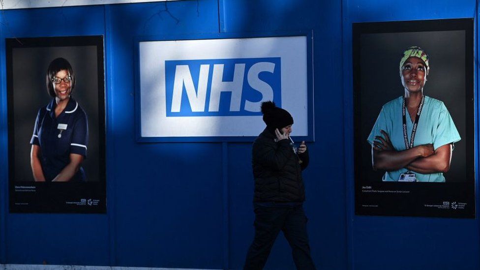 Знак NHS на стене с фотографиями двух работников NHS.