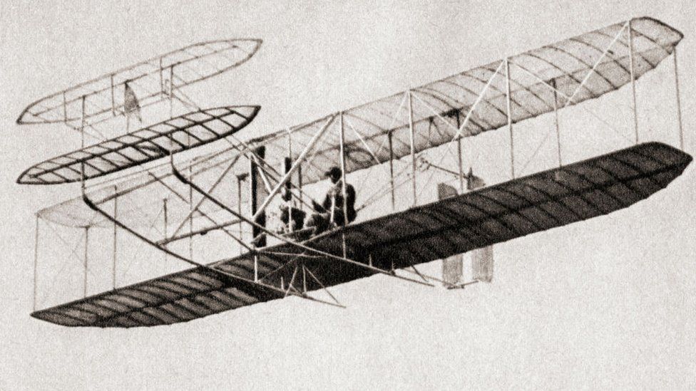 Изображение первого полета братьев Райт, где пилот сидит в кабине и смотрит на лифты, которые двигали самолет вверх и вниз.