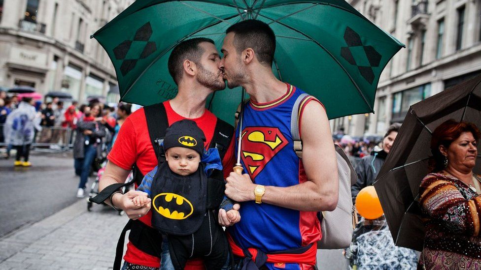 Same-sex couple kiss with one of them holding a baby on his chest