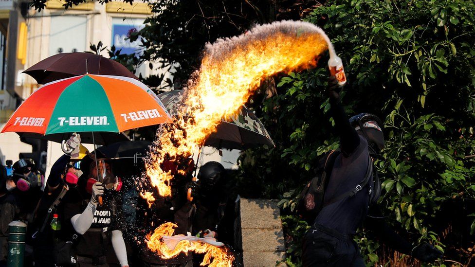 An arc of fire streams from a petrol bomb in a bottle as a black-clad protester tosses it