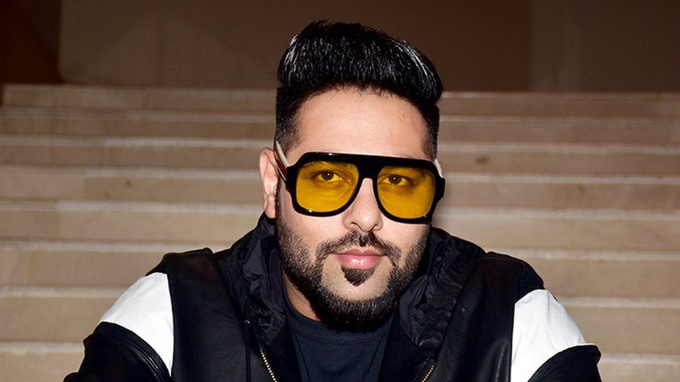 Badshah looking at the camera wearing a black leather jacket with white shoulder pattern. Hee's wearing yellow tinted glasses with his hair slicked up. The background is of light brown coloured long steps.