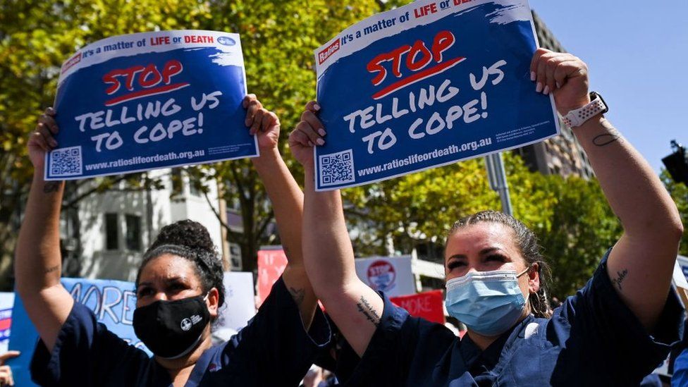 Two healthcare workers at a protest hold a sign saying 'stop telling us to cope'