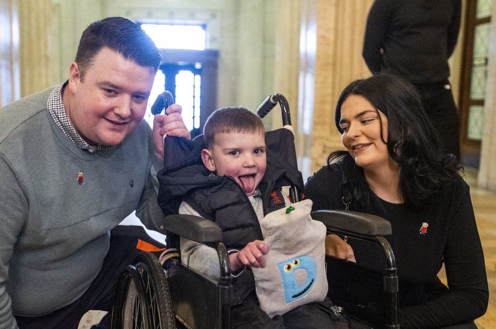 Dáithí Mac Gabhann sits in his wheelchair, holding a hot water bottle as his parents Máirtín and Seph look on