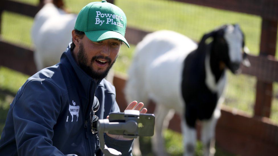 A man holds an iPhone on a stabilising rig while he kneels down next to a goat on his farm