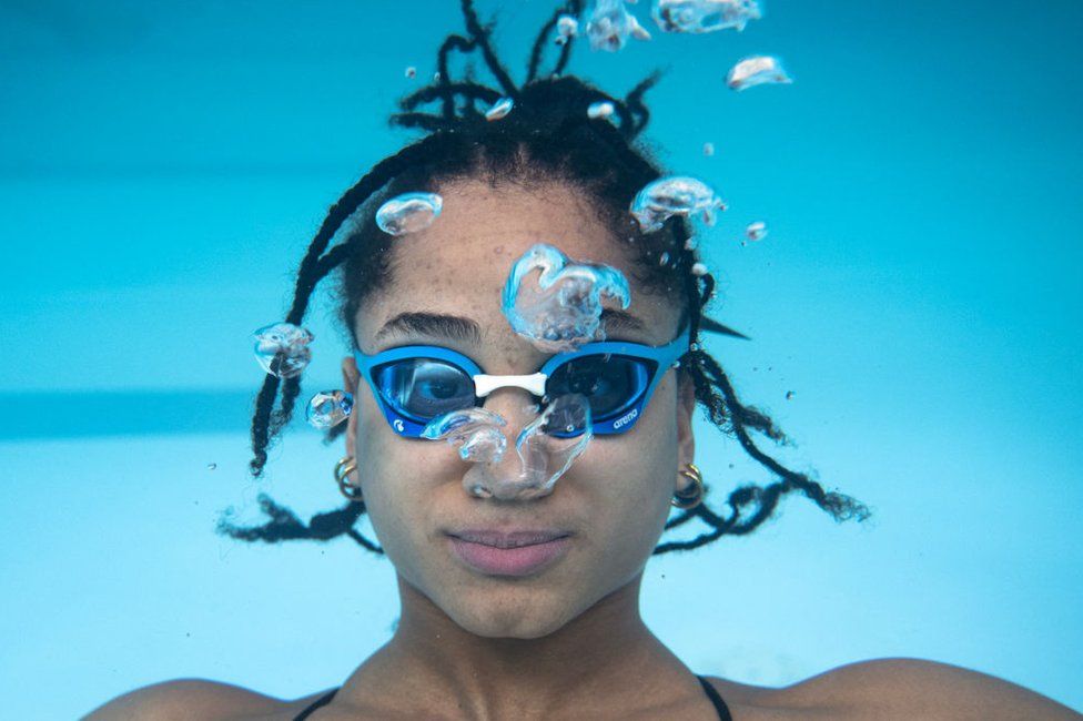 A woman wearing goggles looks at the camera underwater.
