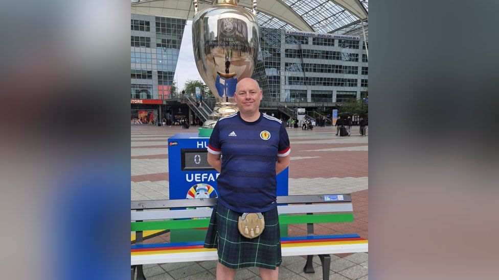 Ross Hamilton wearing a kilt and standing in front of a replica of the Euros trophey