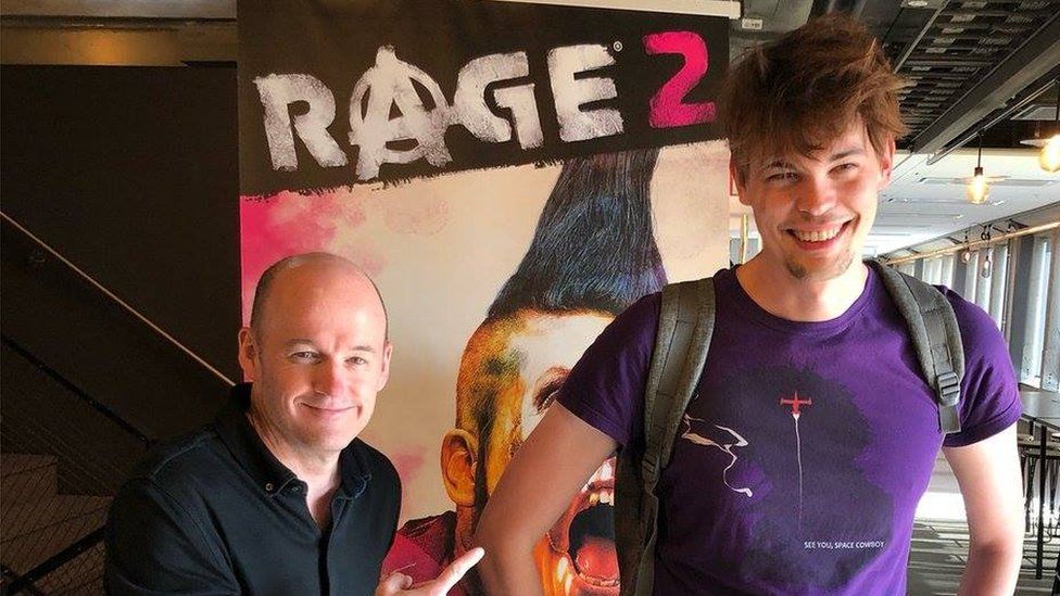 Javy is stands in front of a poster of the first-person shooter game Rage 2