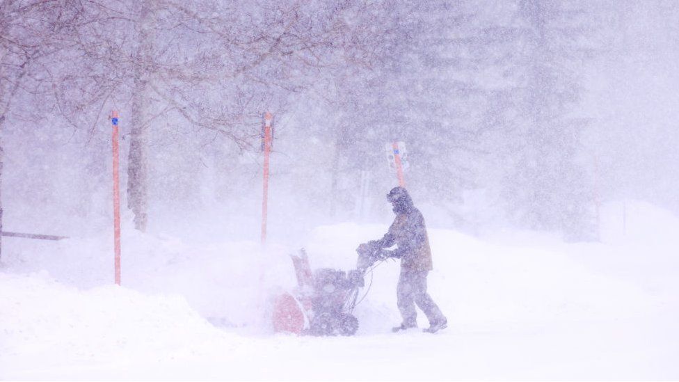 A hooded man with a snow plough tries to remove the snow amid blizzard conditions