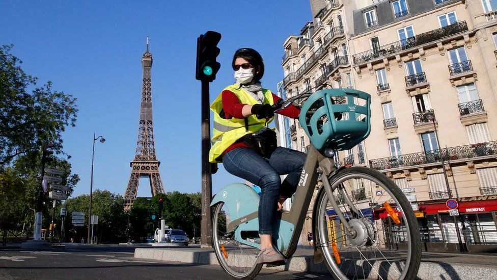 A woman wearing a face mask rides her bicycle in Paris, with the Eiffel Tower captured behind her