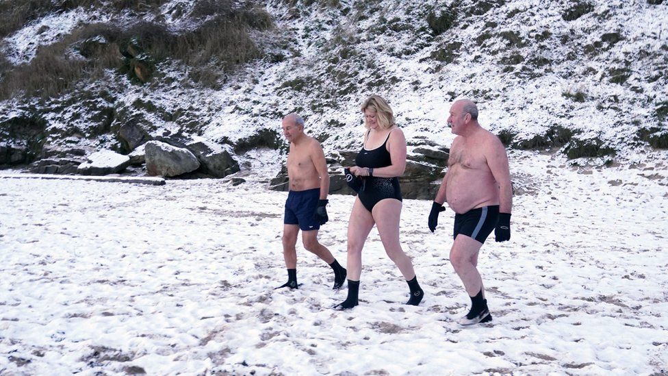 Picture showing two shirtless men and a woman in a swimming costume walking across a field covered in snow
