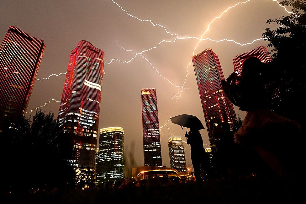 A bolt of lightning crosses the sky as people look at buildings displaying a light show on the eve of the 100th anniversary of the Chinese Communist Party in Beijing
