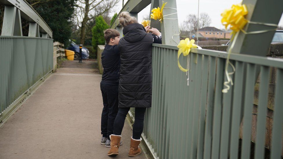 A woman and a young boy tie a yellow ribbon with a message of hope written on it, to a bridge over the River Wyre in St Michael's on Wyre (note - parental permission given)