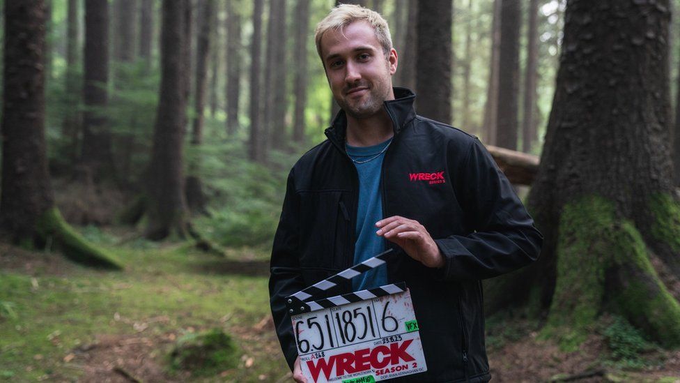 Ryan Brown, pictured in woodland, with a clapperboard for Wreck season two. Ryan is a tall white man in his early 30s with blonde hair cut short and stubble. He wears a black waterproof jacket embroidered with 'Wreck Season 2' and a blue T-shirt.