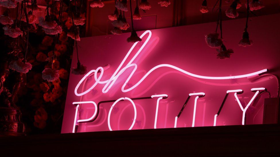 A pink fluorescent sign of the Oh Polly logo against a black backdrop.