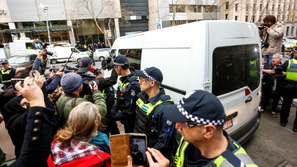A white van carrying George Pell leaves court, beside police officers, media representatives and members of the public