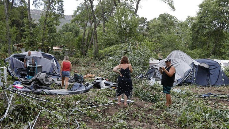 Campers stand among tents destroyed by wind and fallen foliage.