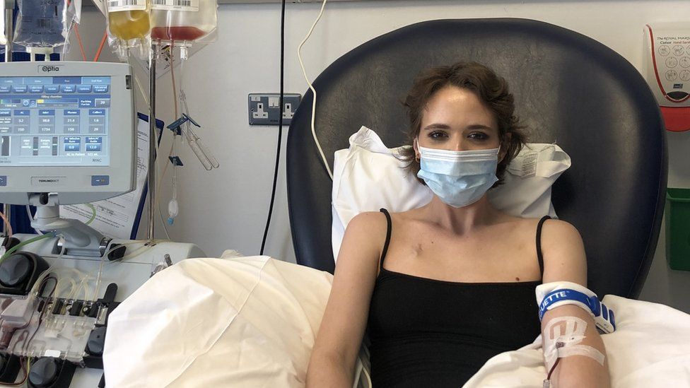 Mattie sitting in hospital bed wearing a black vest top. She has short brown hair, a blue face mask across her face and there are various tubes connected to her arms. There is a hospital machine to the left of her in bed.